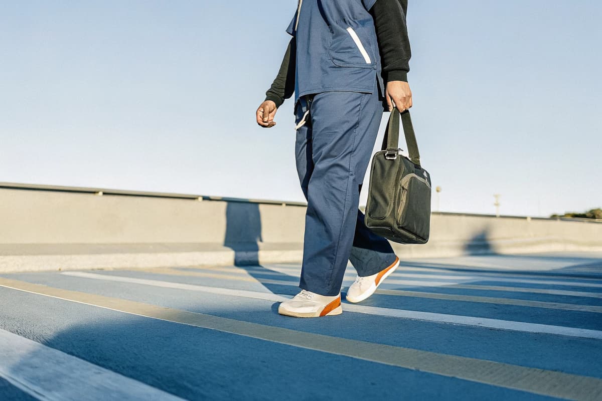 Healthcare worker in navy scrubs walking through a hospital parking garage at golden hour, wearing white sneakers with orange accents and a black hoodie draped over one shoulder