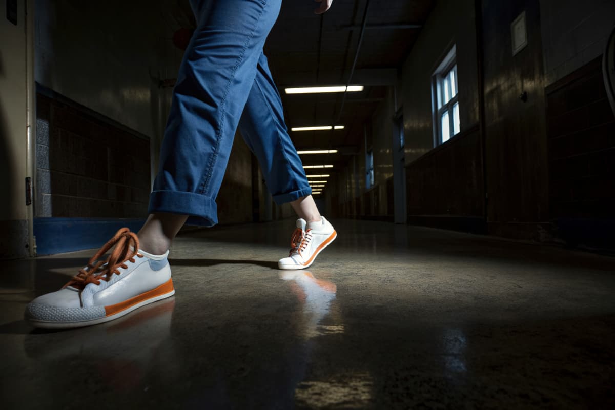 Close-up of a nurse walking through a hospital hallway in navy scrubs and white sneakers with orange sole accents and orange laces, low angle, fluorescent lighting, face out of frame