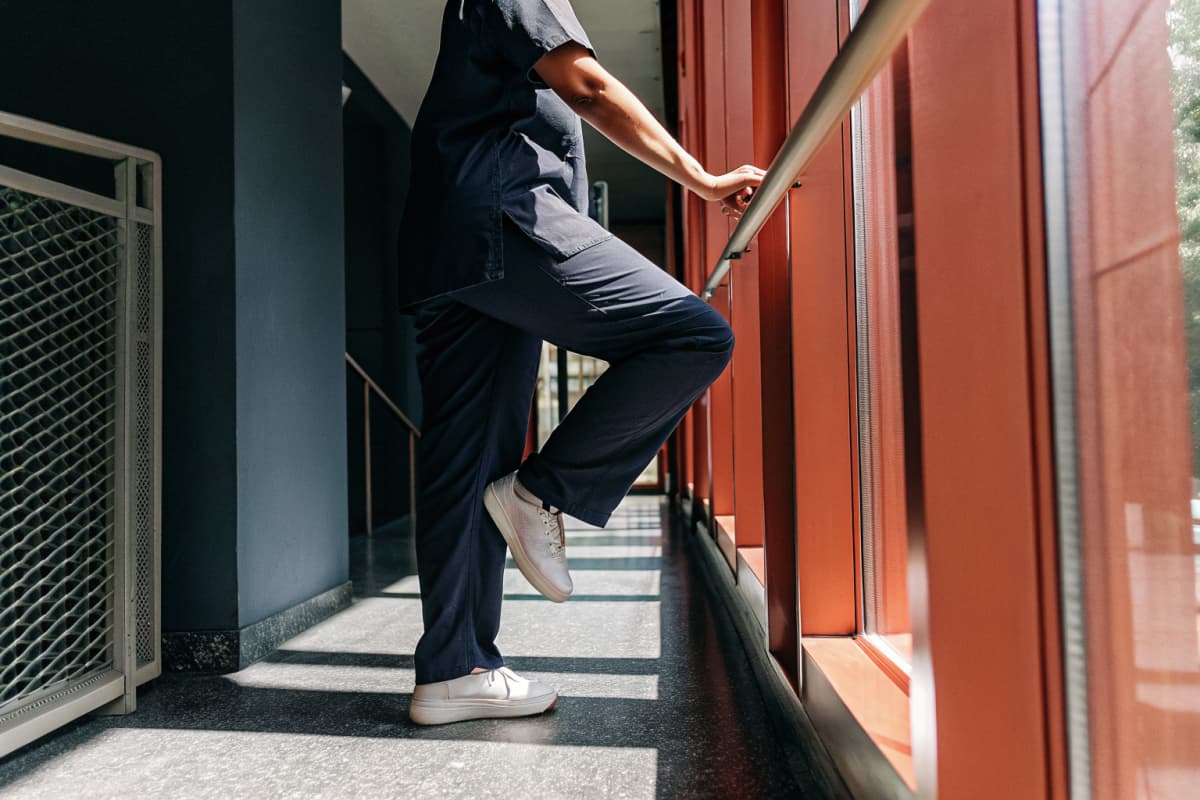 Nurse in navy scrubs doing a standing calf raise stretch against a rail in a hospital hallway, white sneakers visible on her feet