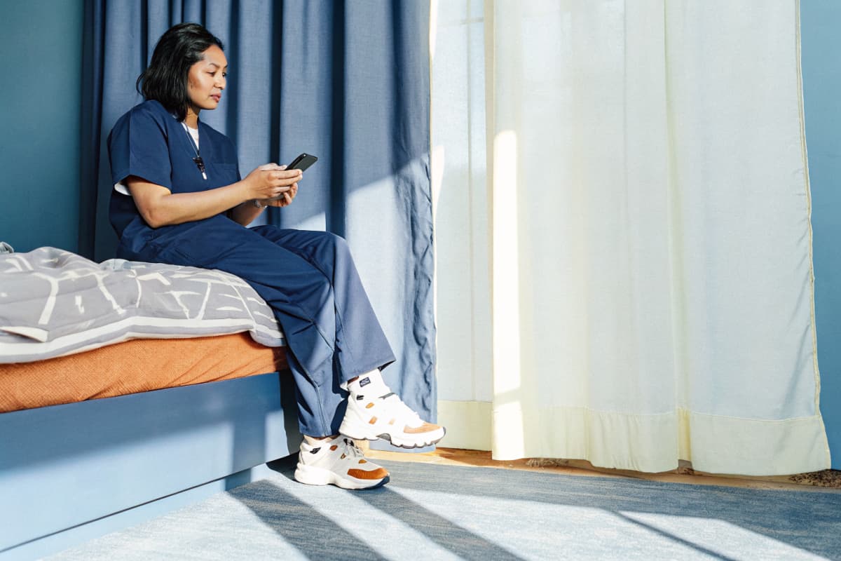 Nurse in navy scrubs sitting on the edge of a bed with blackout curtains drawn behind her, checking a sleep tracking app on her phone, white sneakers with orange accents on the floor beside the bed
