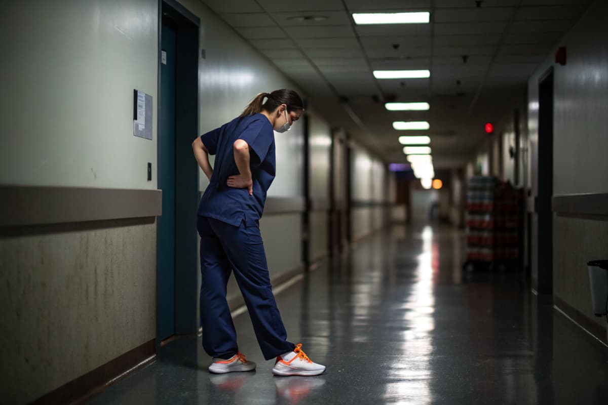 Nurse in navy scrubs standing alone in a dim hospital corridor at the end of a shift, one hand on her lower back, white and orange sneakers visible