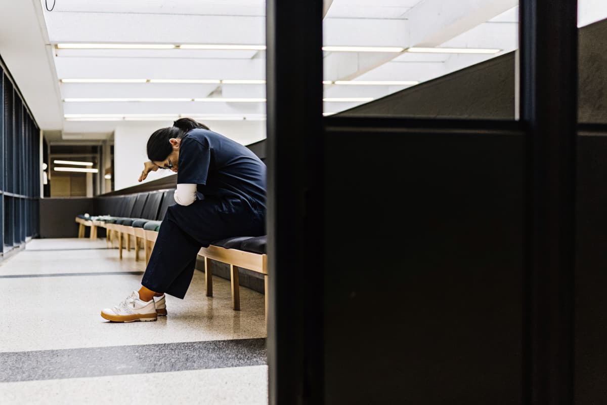 Nurse in navy scrubs sitting alone on a hospital bench, head down, white and orange sneakers on her feet