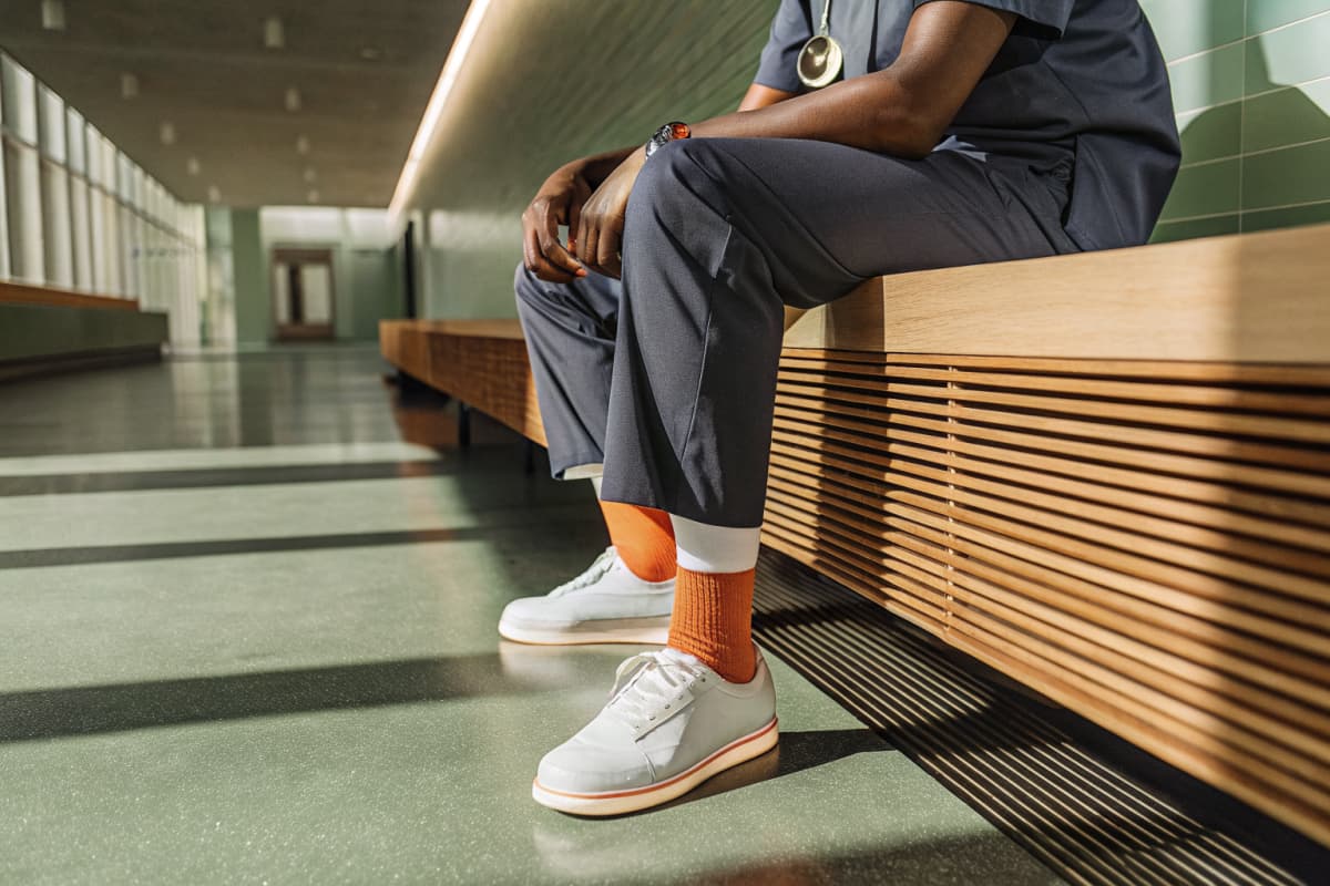 Nurse in navy scrubs sitting on a bench in a bright hospital hallway during a break, hands clasped, head down, white sneakers with orange accents and orange compression socks visible