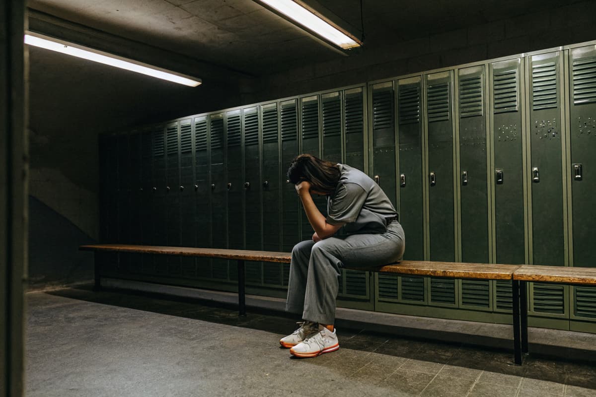 Nurse in grey scrubs sitting alone on a locker room bench, head down resting on her hand, white and orange sneakers visible, exhausted after a shift