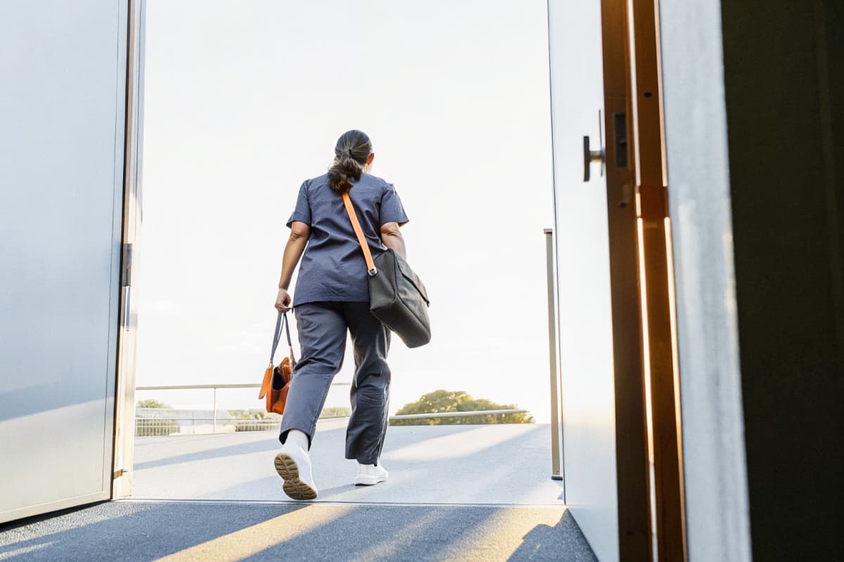Nurse in navy scrubs seen from behind walking out of a hospital exit into golden afternoon light, orange lanyard badge reel and gym bag over one shoulder, white sneakers visible on the pavement, heading into off-shift hours