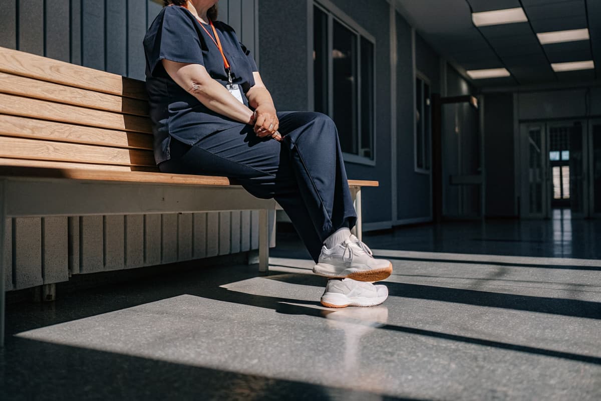 Nurse in navy scrubs sitting on a break room bench in warm window light, orange badge reel lanyard resting on her lap, white sneakers with orange sole accents visible on the floor, face out of frame