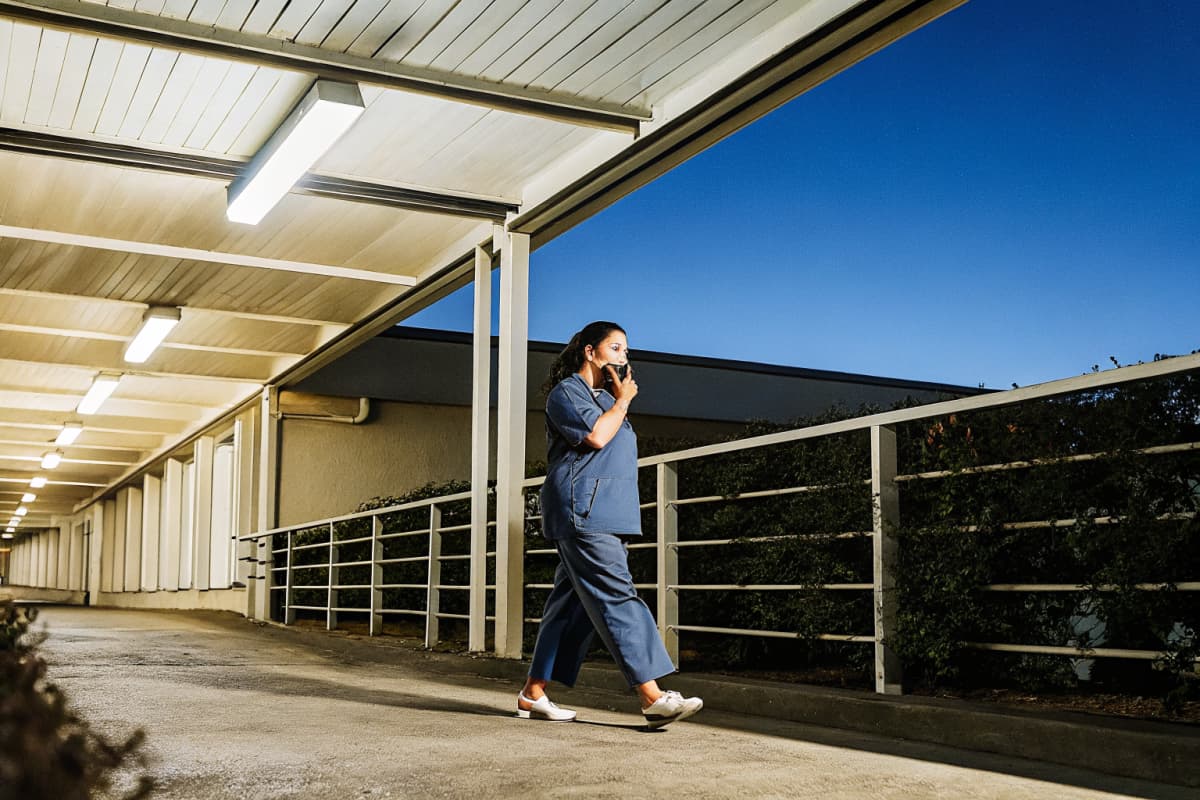 Nurse in navy scrubs and white sneakers walking alone down a covered hospital walkway at night while checking her phone