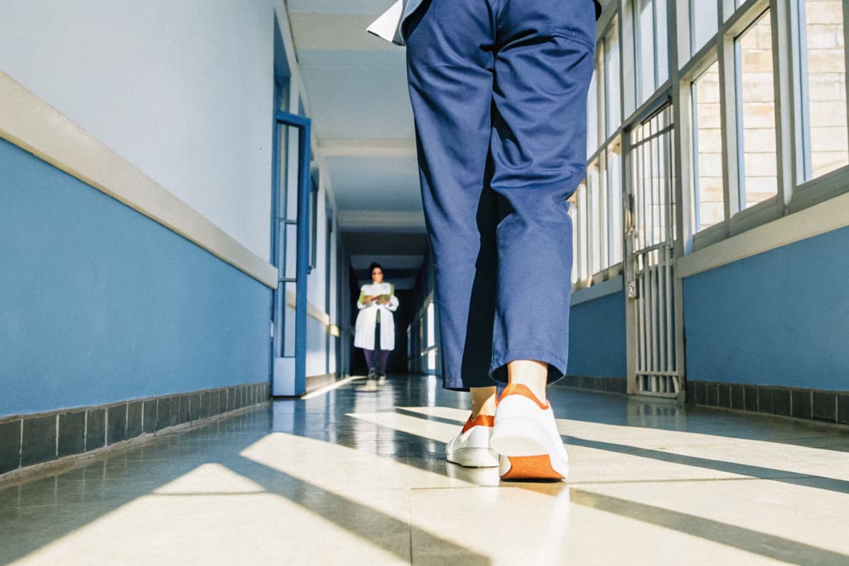Low-angle shot of a nurse in navy scrubs and orange and white sneakers walking down a bright hospital hallway with a hospital administrator observing in the background
