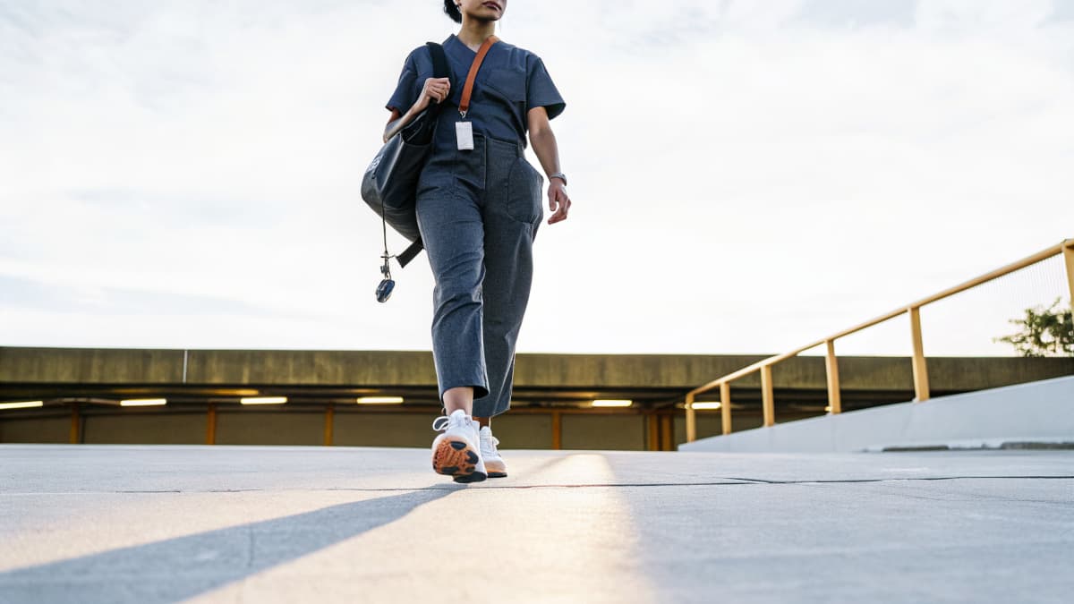 Nurse in scrubs walking through a hospital parking garage at dusk, ID badge on an orange lanyard swinging at her waist, white sneakers with orange sole accents visible on the pavement