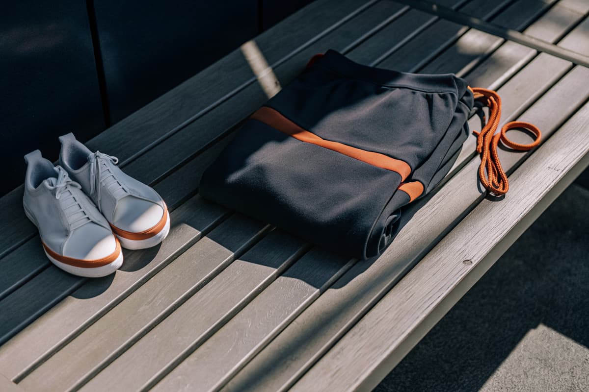 Navy and orange colorblock hoodie folded neatly next to a matching pair of white sneakers with orange sole accents and a coiled orange lanyard, on a sunlit locker room bench