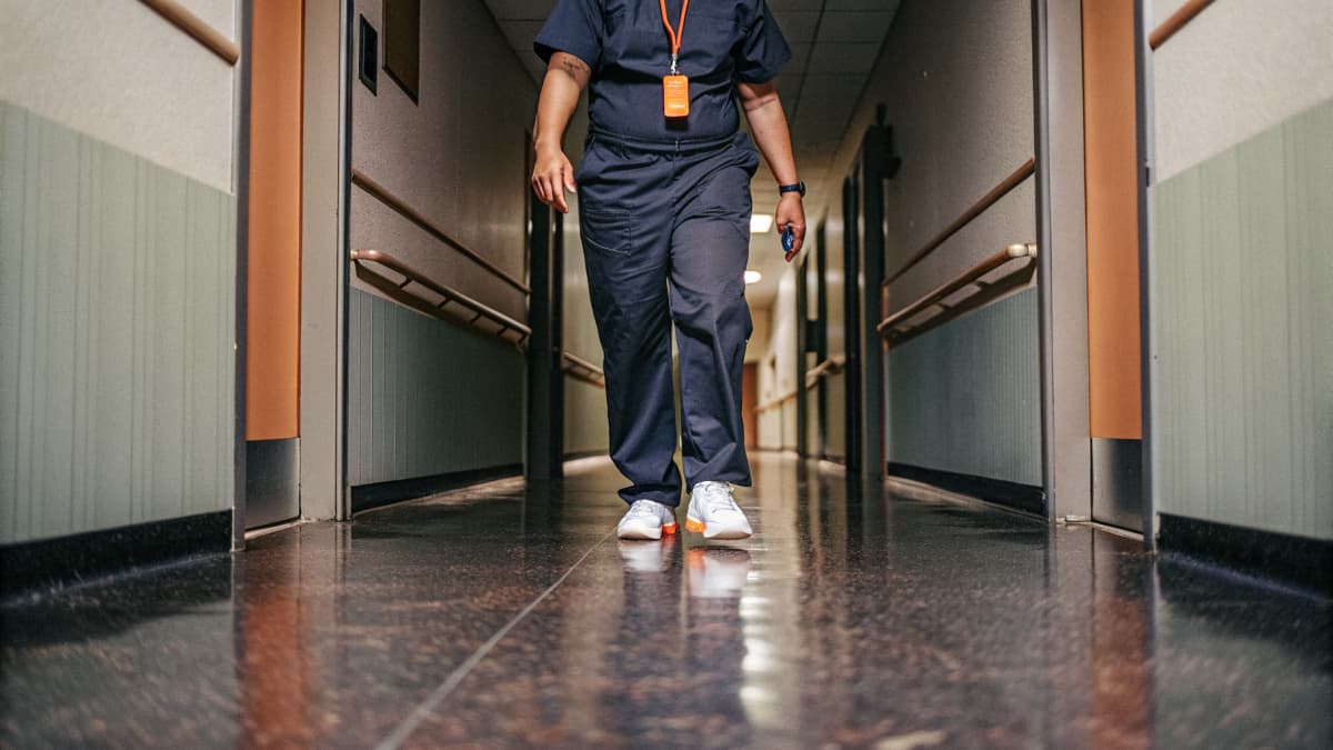 New grad RN in navy scrubs with a badge reel clipped to her waistband, white and orange sneakers visible, walking down a bright hospital hallway at shift change