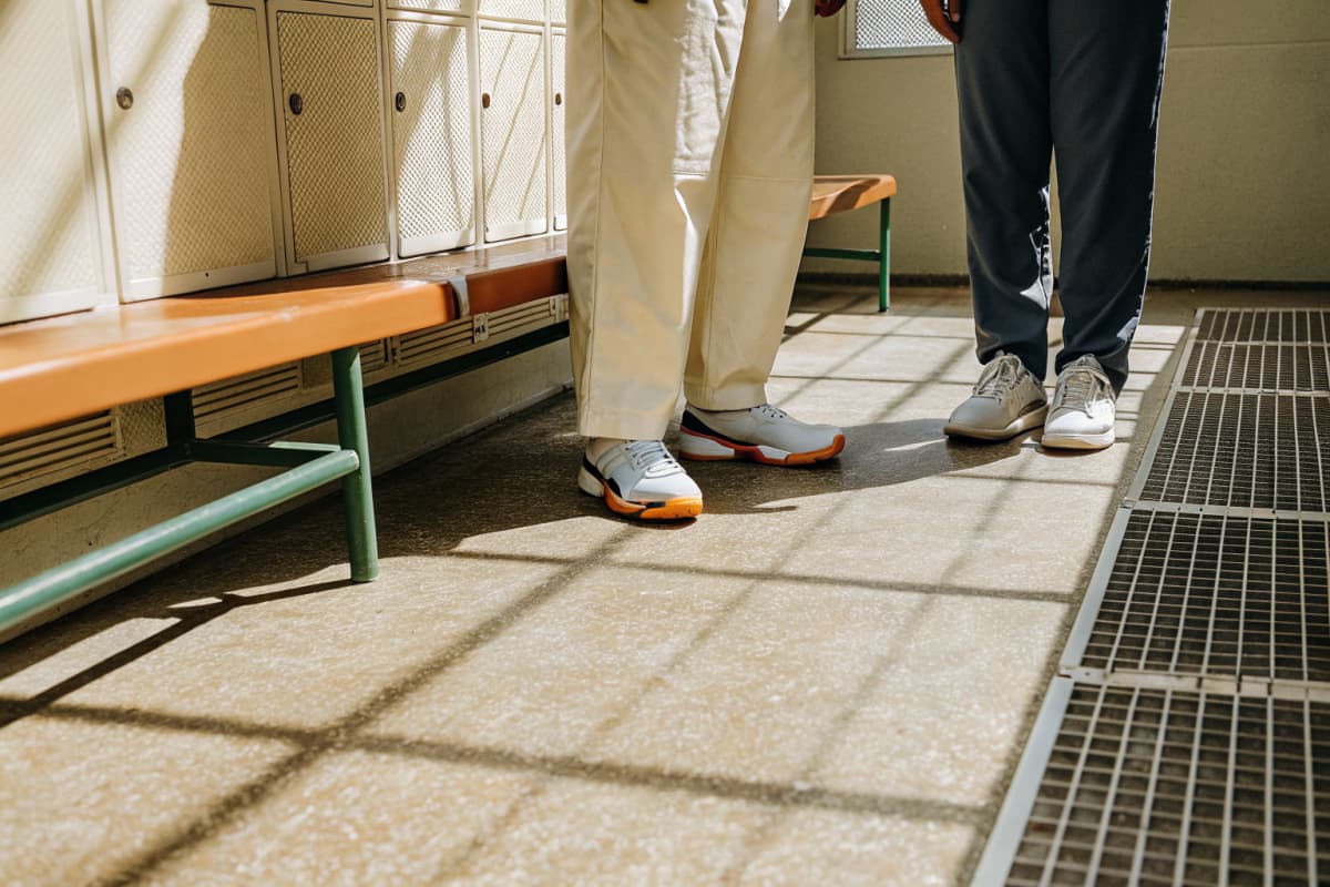 Two healthcare workers standing in a hospital locker room, cropped at the waist, wearing scrub pants and clean white sneakers with orange sole accents on sunlit tile floor