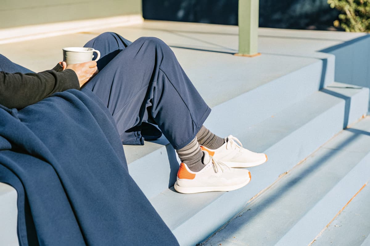Healthcare worker in navy scrubs relaxing on porch steps during golden hour on a day off, holding a mug, wearing white sneakers with orange accents and a black hoodie draped over their lap
