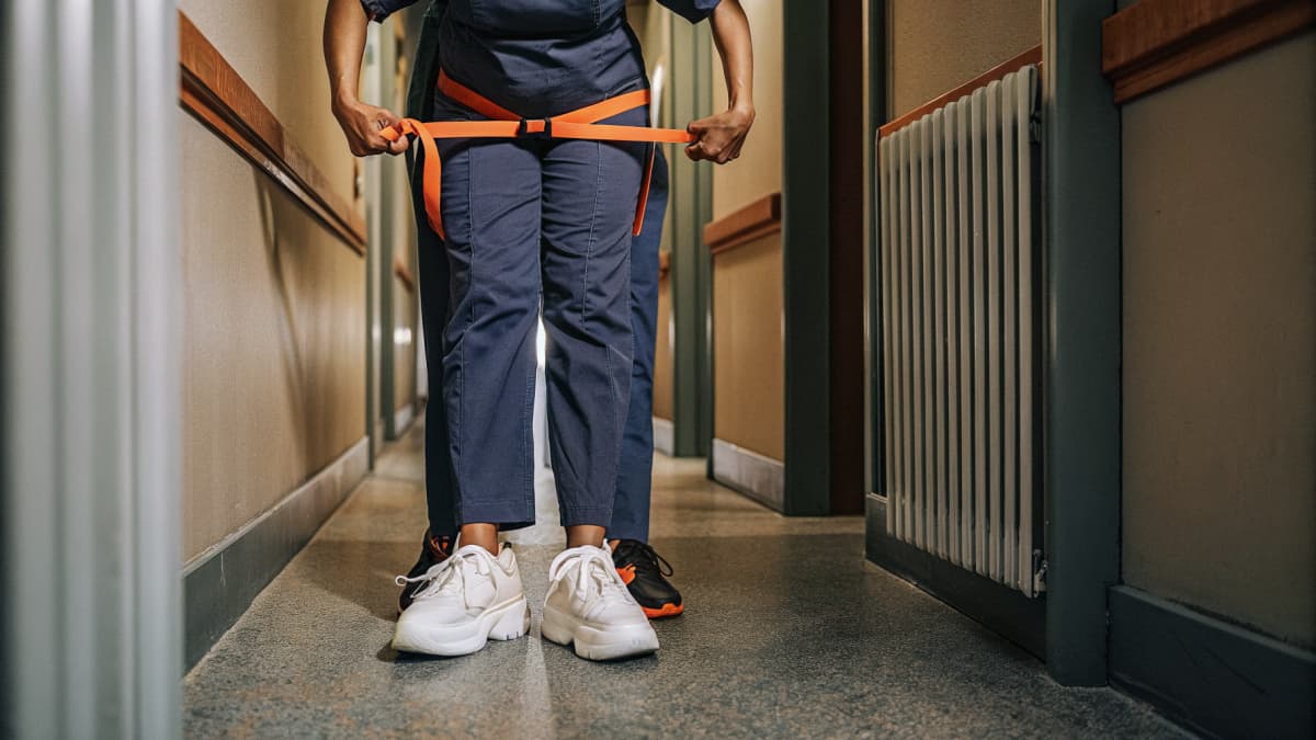 Nurse in navy scrubs bracing a gait belt around a patient in a hospital hallway, white sneakers with orange accents planted in a stable stance