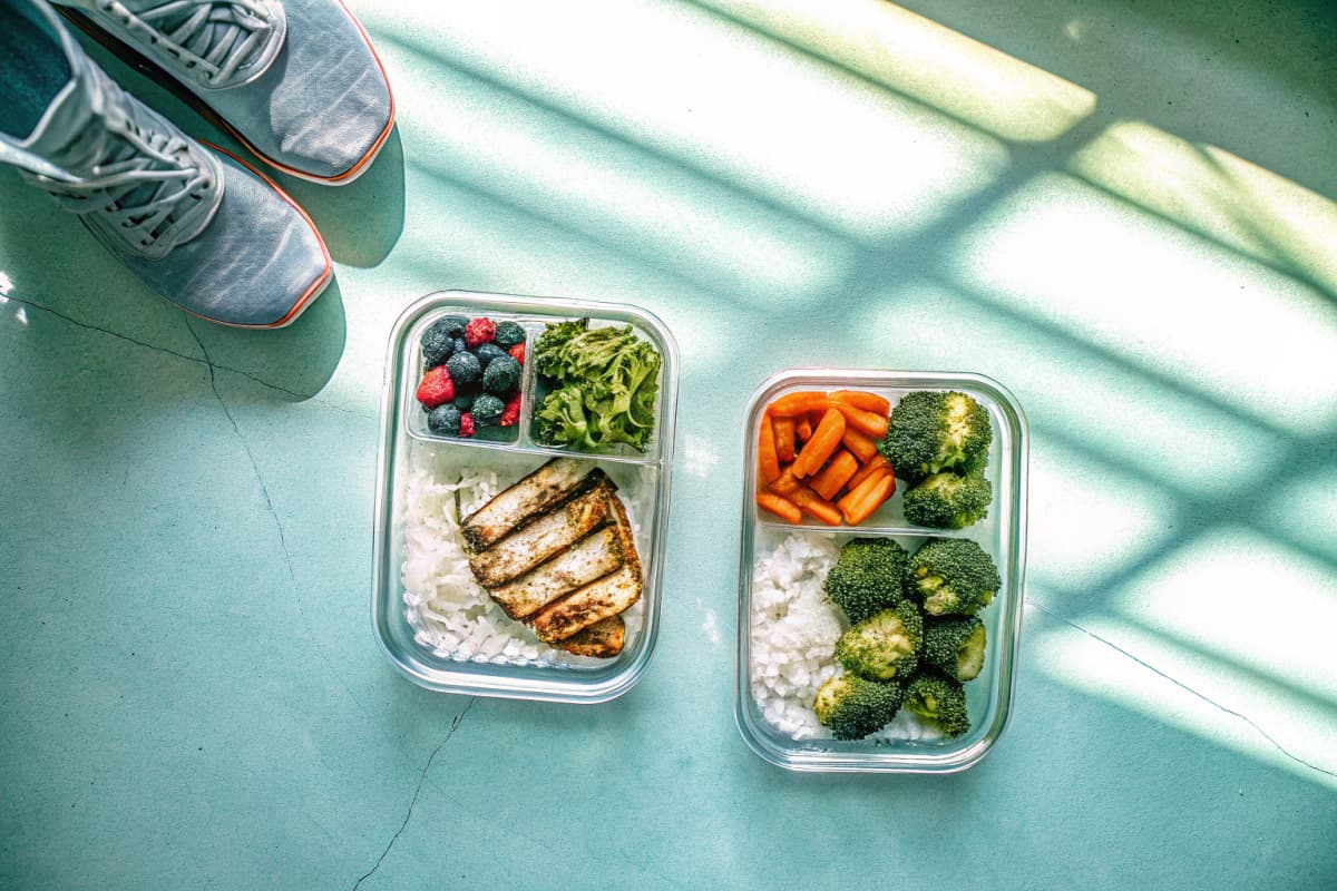 Overhead flat-lay of two glass meal-prep containers packed with grilled chicken, rice, roasted vegetables, and berries next to a pair of sneakers on a countertop