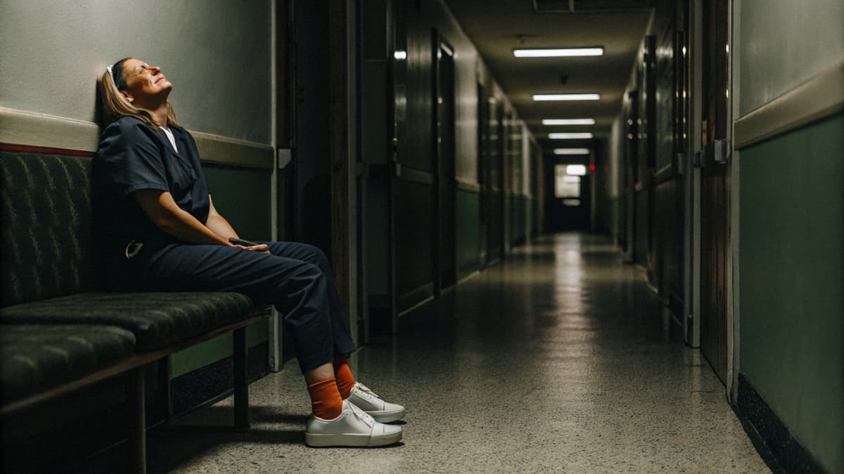 Exhausted nurse in navy scrubs sitting alone on a hospital hallway bench, head back against the wall, white sneakers with orange accents visible on her feet