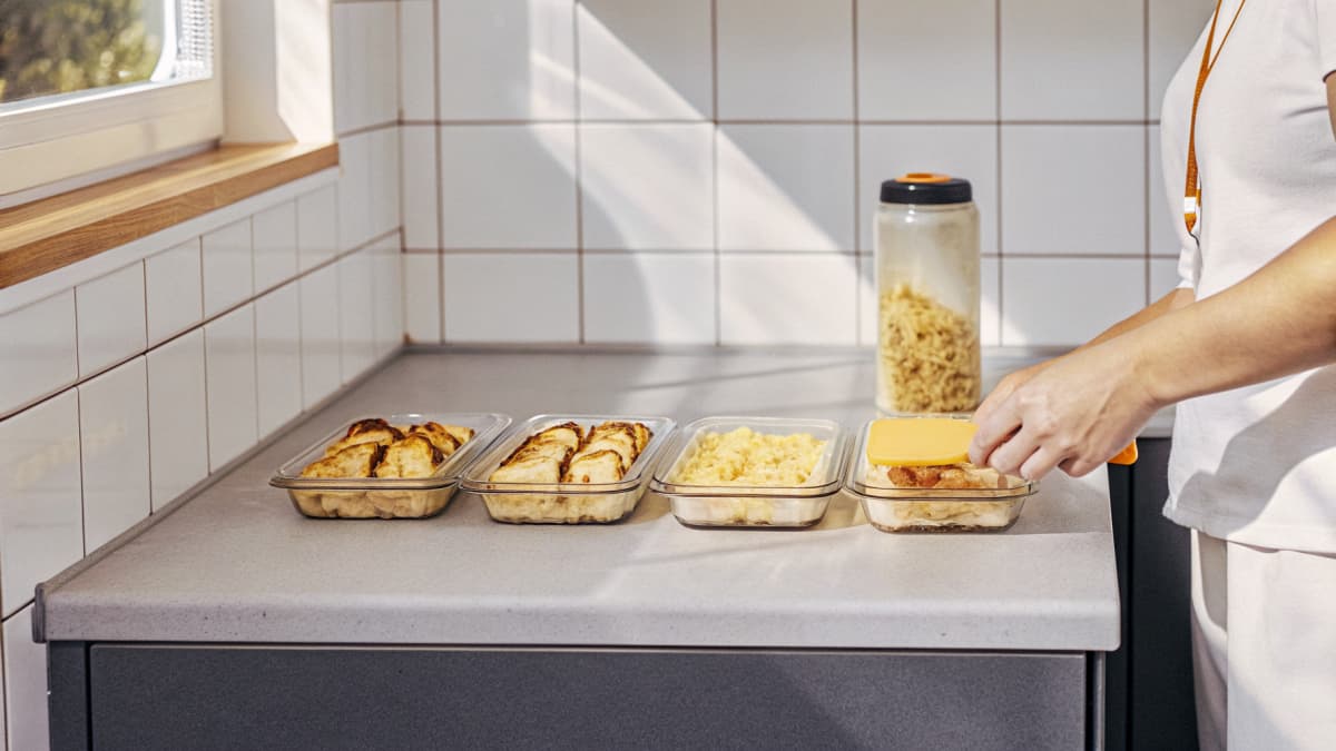 Close-up of a nurse's hands packing four glass meal-prep containers with grilled chicken, rice, and roasted vegetables on a kitchen counter, orange badge reel resting nearby, white sneakers with orange sole accents visible on the floor, face out of frame