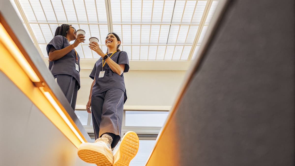 Two healthcare workers in navy scrubs sharing coffee cups and laughing together, orange badge reel lanyards visible, white sneakers with orange sole accents in frame, hospital setting