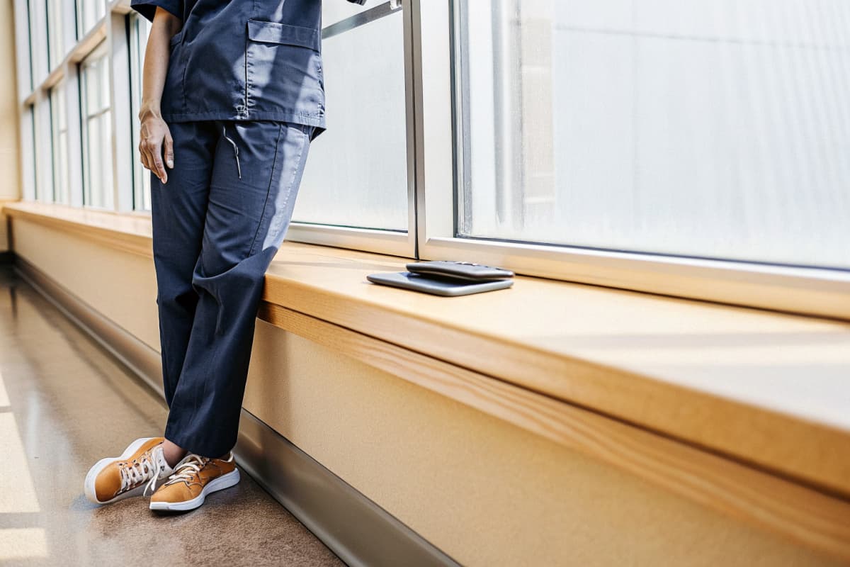 Nurse in navy scrubs and orange sneakers standing beside a sunlit hospital window during a quiet break, phone resting on the windowsill