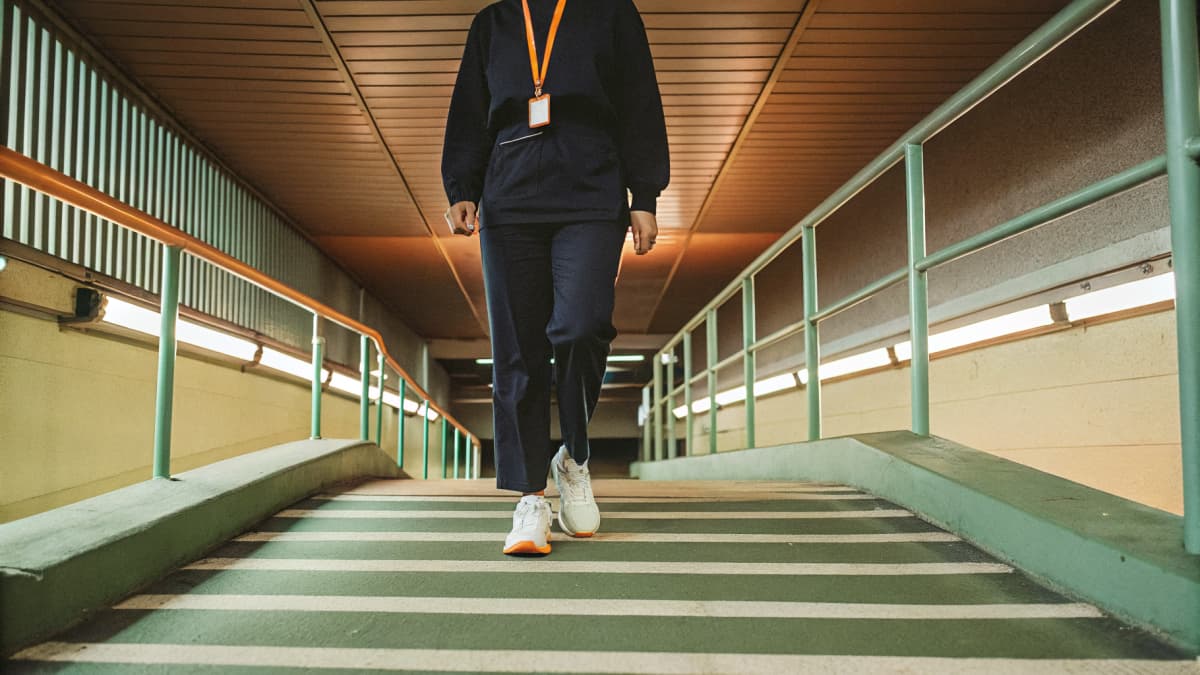 Nurse in navy scrubs walking through a hospital parking garage at dawn, white sneakers with orange accents visible, black hoodie over one shoulder