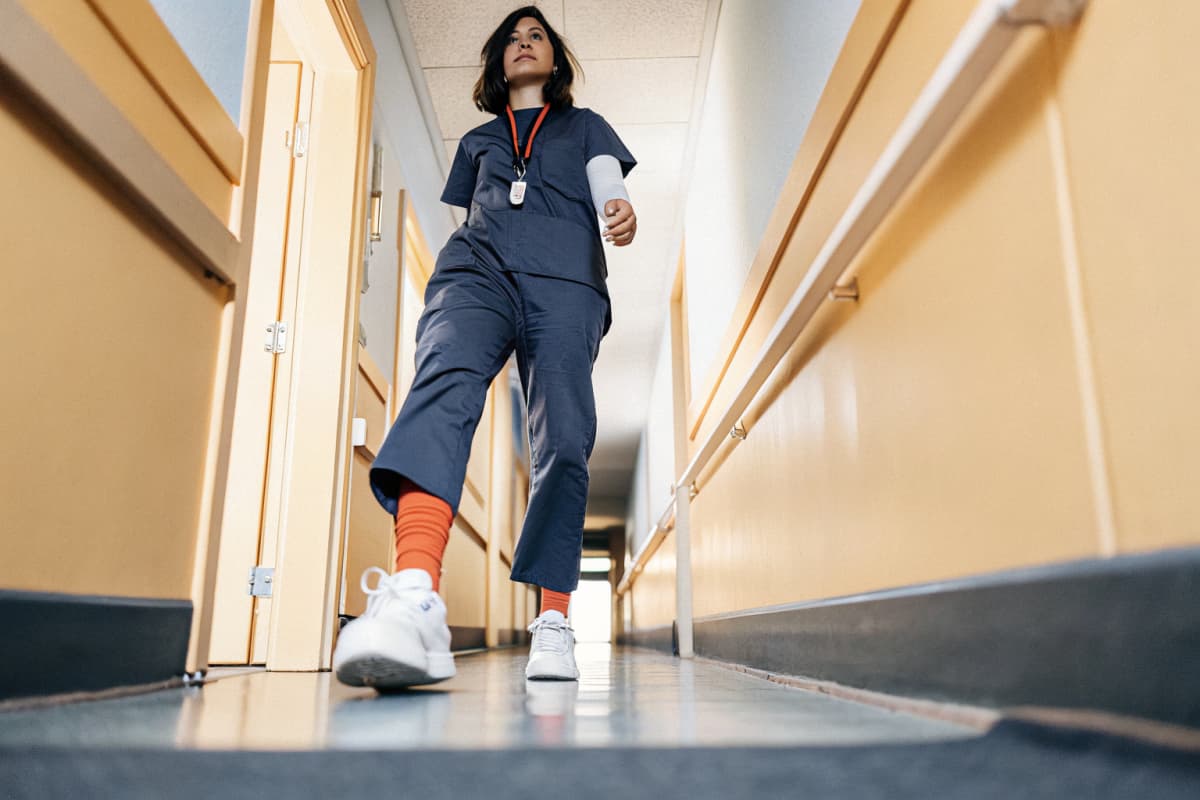 New nurse in navy scrubs walking down a hospital hallway on her first shift, orange compression socks and white sneakers with orange accents visible, badge reel around her neck