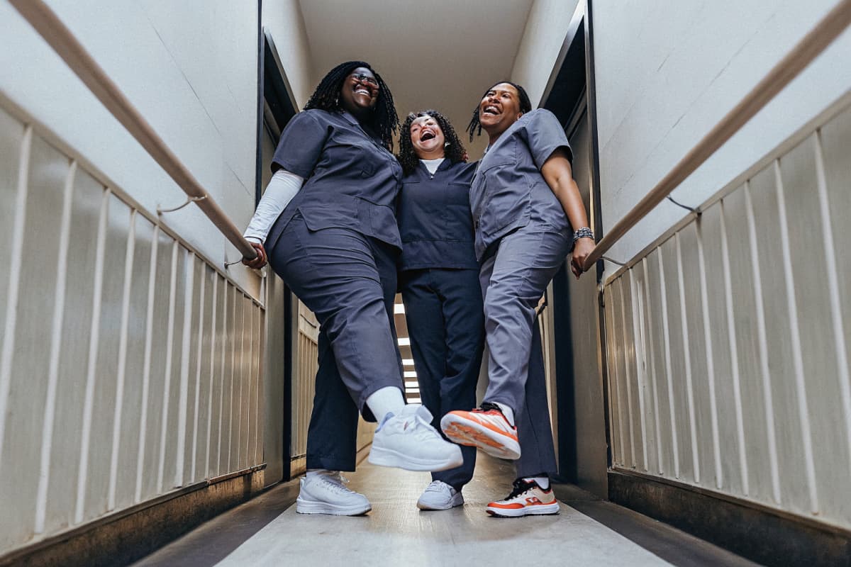 Three nurses in scrubs laughing together in a hospital hallway during shift change, white and orange sneakers visible