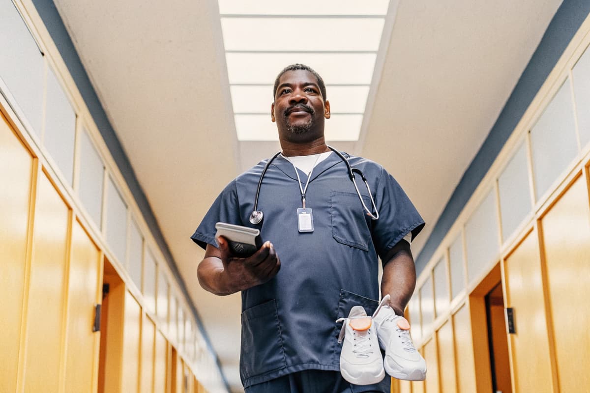Proud male RN in navy scrubs standing in a bright hospital hallway holding a certification card, stethoscope around his neck, white sneakers with orange accents visible