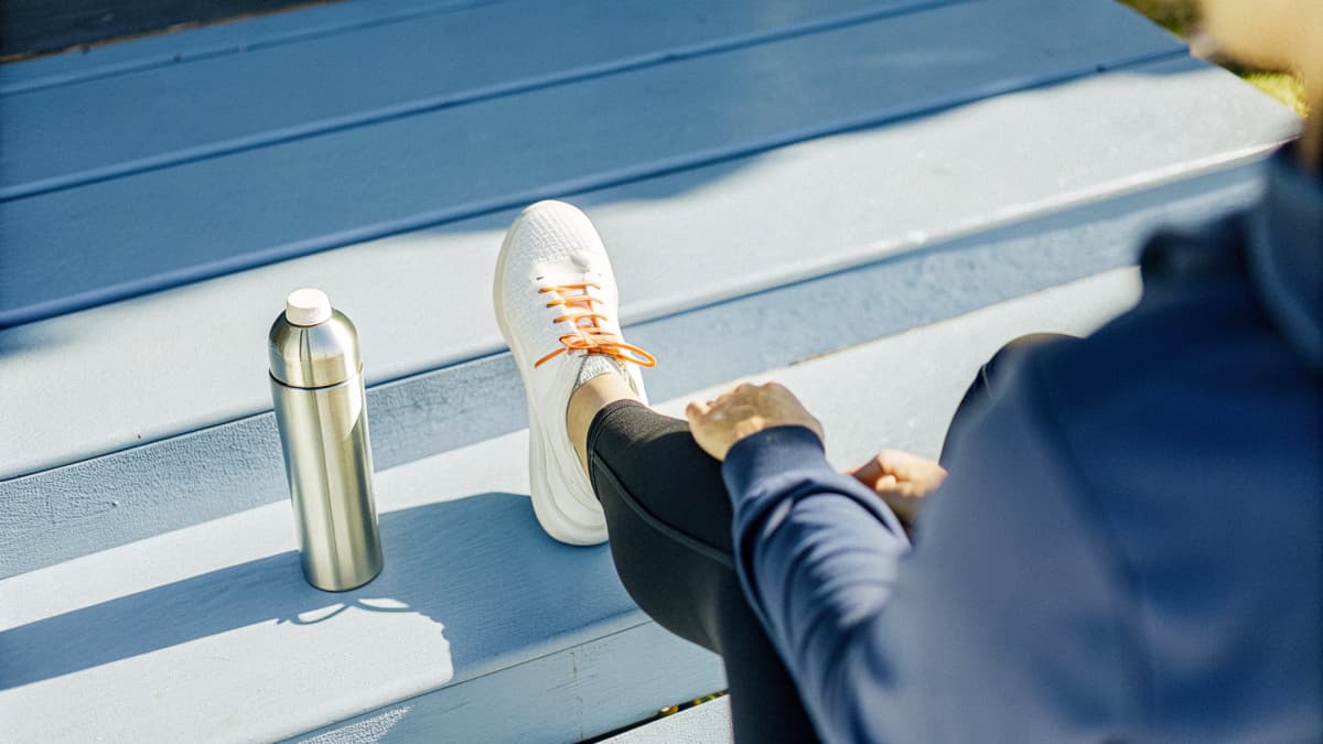 Healthcare worker seen from behind on porch steps in morning light, lacing up a white sneaker with orange sole accents and orange laces, wearing a navy hoodie and black joggers, a stainless water bottle and rolled yoga mat resting nearby, face out of frame