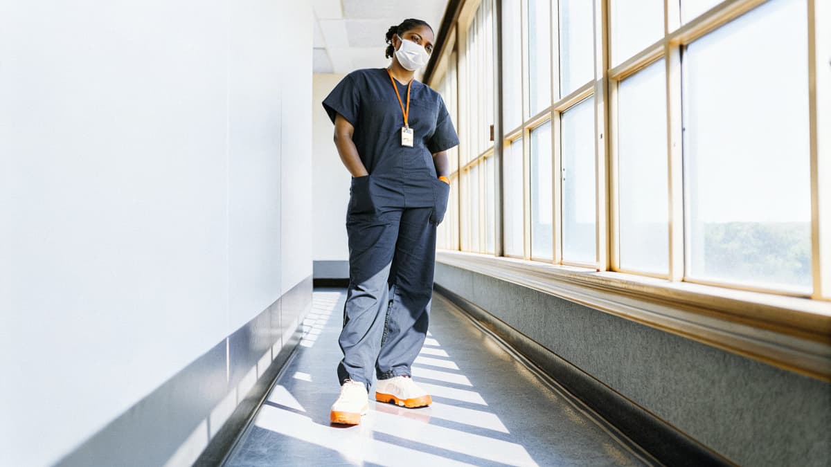 Nurse in navy scrubs standing confidently in a sunlit hospital hallway, orange badge lanyard, white sneakers with vivid orange sole accents visible on her feet, face turned away for privacy