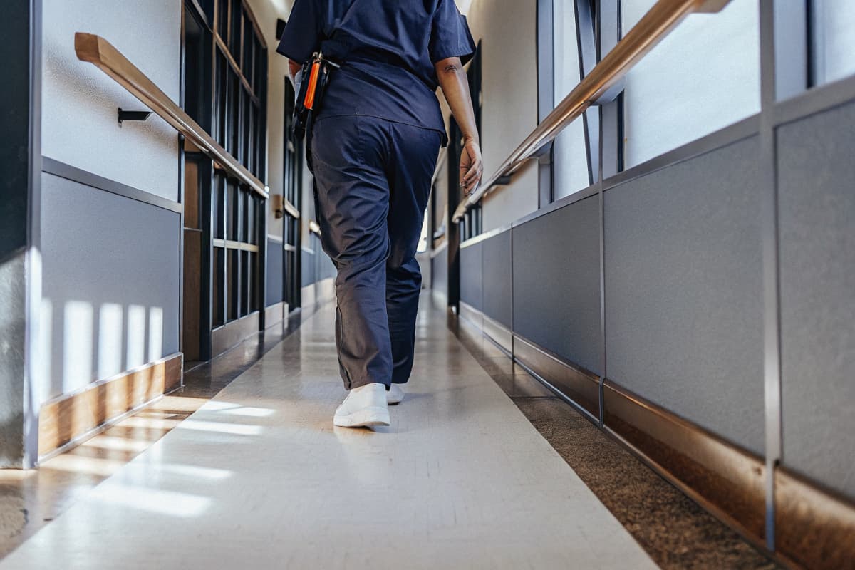 Nurse in navy scrubs and white sneakers with an orange badge reel walking away down an empty hospital hallway